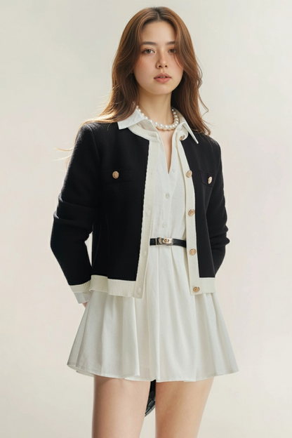 一位女士穿著 the.lookbook.select 的 Moiselle 襯衫連衣裙，搭配黑色開襟衫、珍珠項鍊和黑色腰帶，在素淨的淺色背景下，面朝前方。.