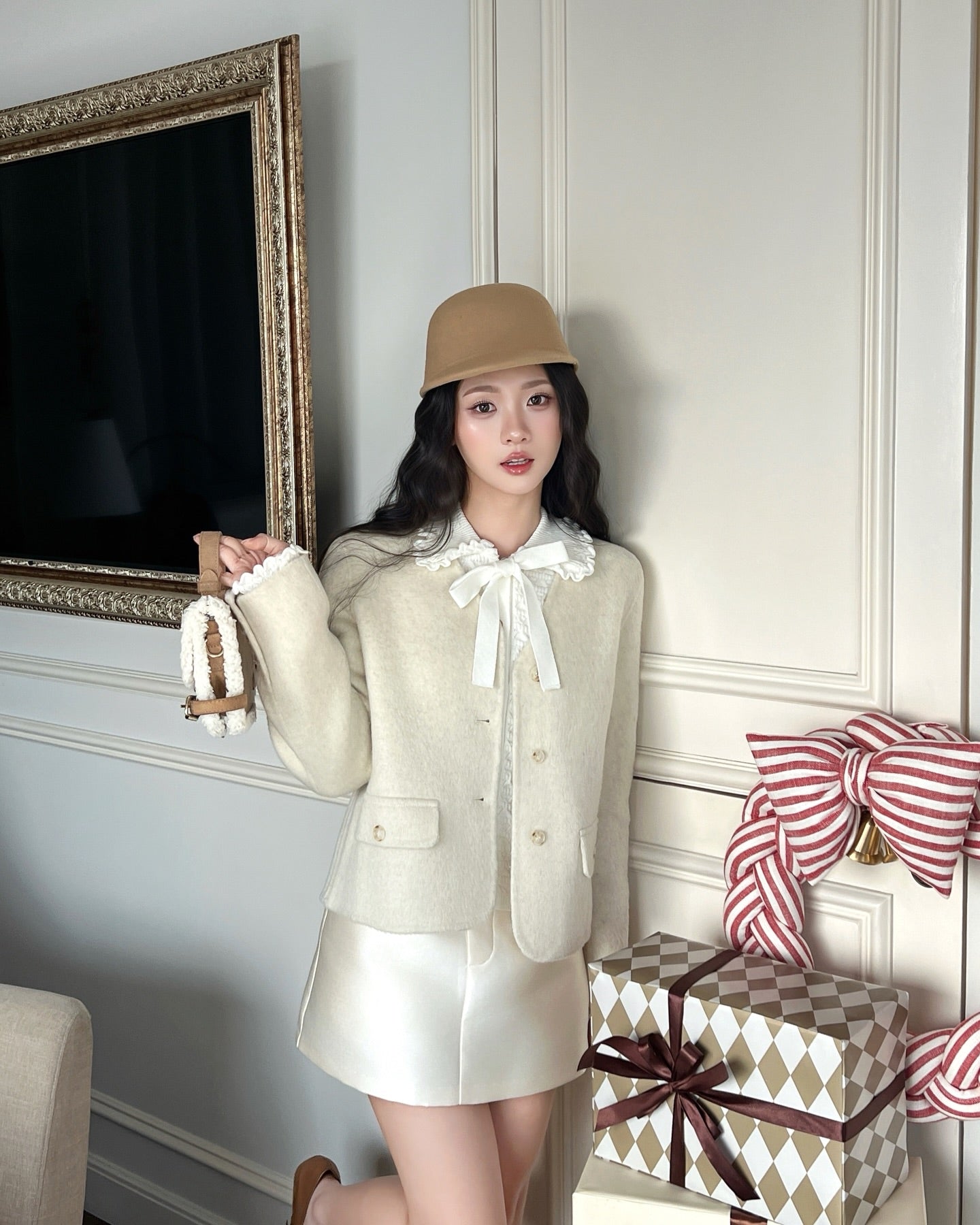 一位穿著 the.lookbook.select's Snow Wool Coat❄️，無領設計，白色短裙和棕褐色帽子的女士站在室內包裝好的禮物和一個大蝴蝶結旁。.