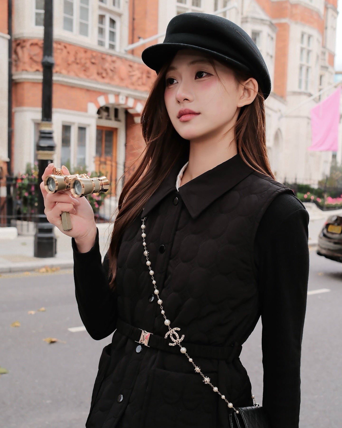 身著 the.lookbook.select 的 Diana Vest Coat，戴著貝雷帽的黑衣女子站在城市街道上，散發著法國精緻風格；她手持小型望遠鏡，背著 Chanel 手袋。.