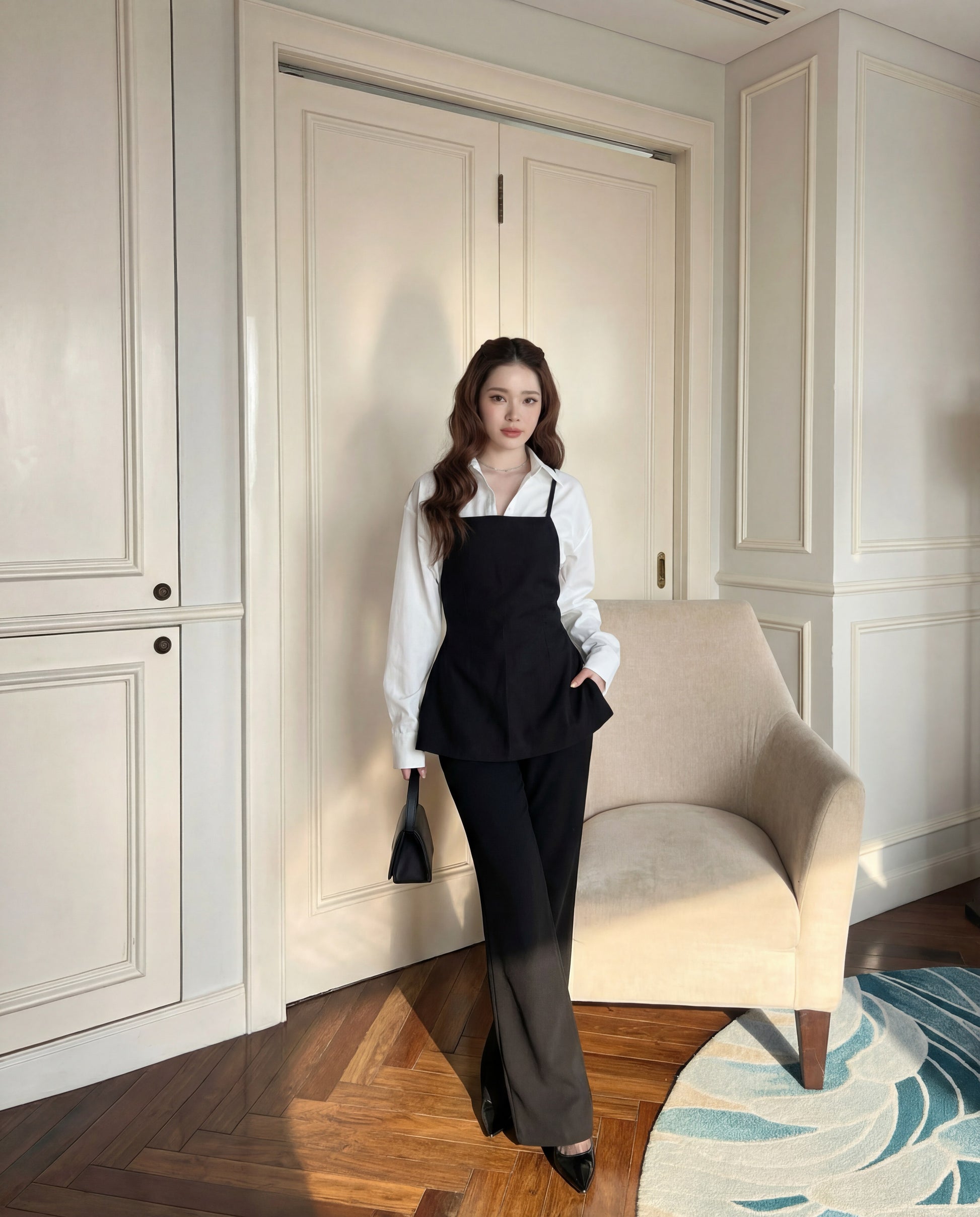 一位室內女性站在米色扶手椅旁，身穿 the.lookbook.select 的 Moet Pants Set 白色襯衫，下穿赫本風格的吊帶背心，搭配寬腳西裝褲和黑色皮鞋，手拿黑色小包。.