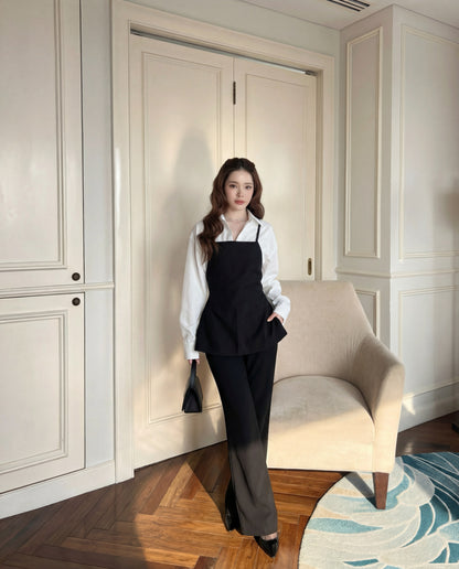 一位室內女性站在米色扶手椅旁，身穿 the.lookbook.select 的 Moet Pants Set 白色襯衫，下穿赫本風格的吊帶背心，搭配寬腳西裝褲和黑色皮鞋，手拿黑色小包。.