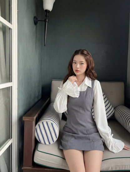 一位留著長髮的年輕女性坐在窗邊的軟墊長椅上，身穿 the.lookbook.select 的灰色 Junior 無袖連身裙，外搭一件荷葉邊袖的白色上衣。.
